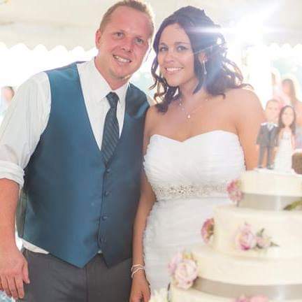 Megan and Brandon Standing next to their 3 teir wedding cake with bobble heads of themselves as cake toppers inside a tent